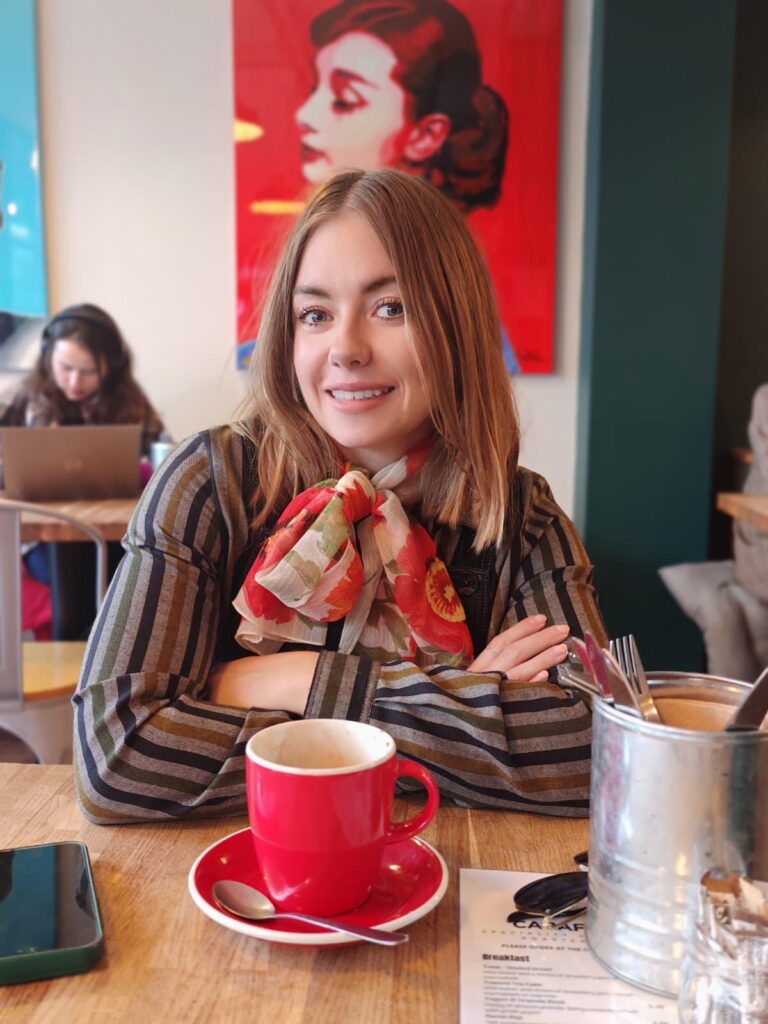 Eleanor Gibson at a café table, arms folded, a red poppy-print chiffon scarf tied at her neck