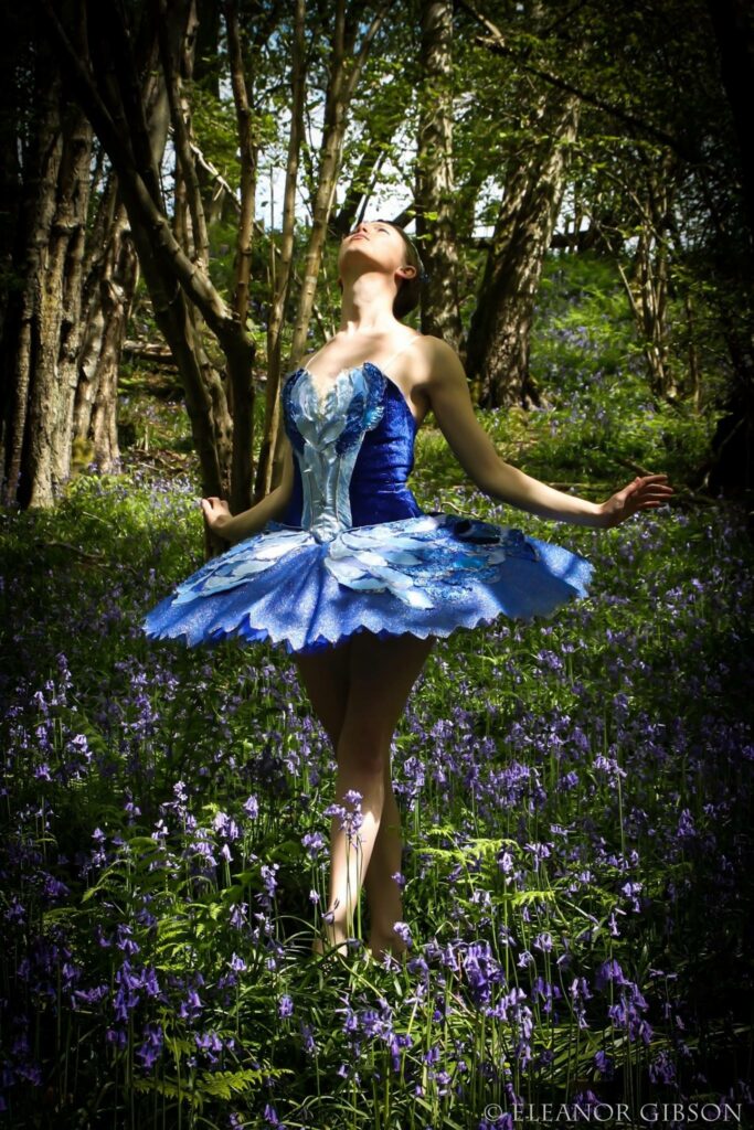 A dancer in a royal-blue and silver tutu, arms open, standing among bluebells in a wood
