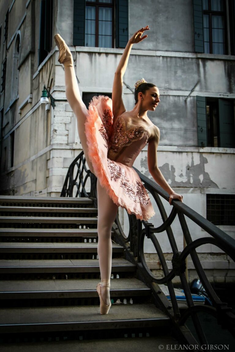 A dancer in a peach tutu with a beaded bodice, one leg raised high, beside the iron railing of a bridge in Venice