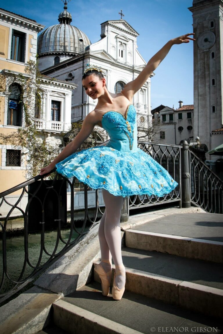 A dancer in a turquoise tutu with an embroidered bodice, one arm raised, on the steps of a bridge in Venice, a domed church behind her