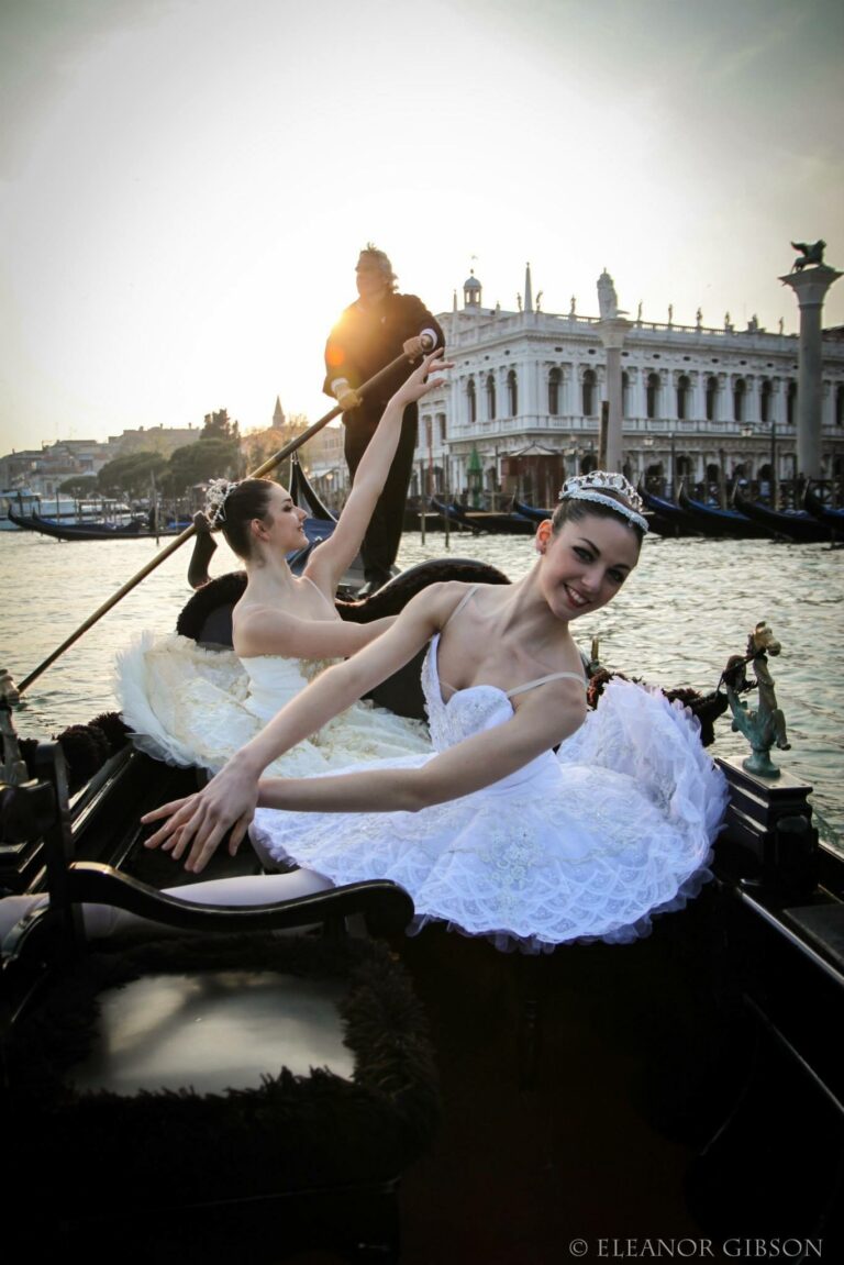 Two dancers in white tutus and tiaras sitting in a gondola on the water in Venice, the gondolier standing behind them